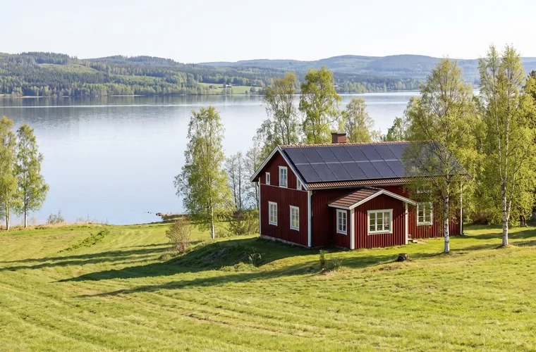Rött timmerhus i Dalarna med solceller på taket, sjö och skogsklädda höjder bakom